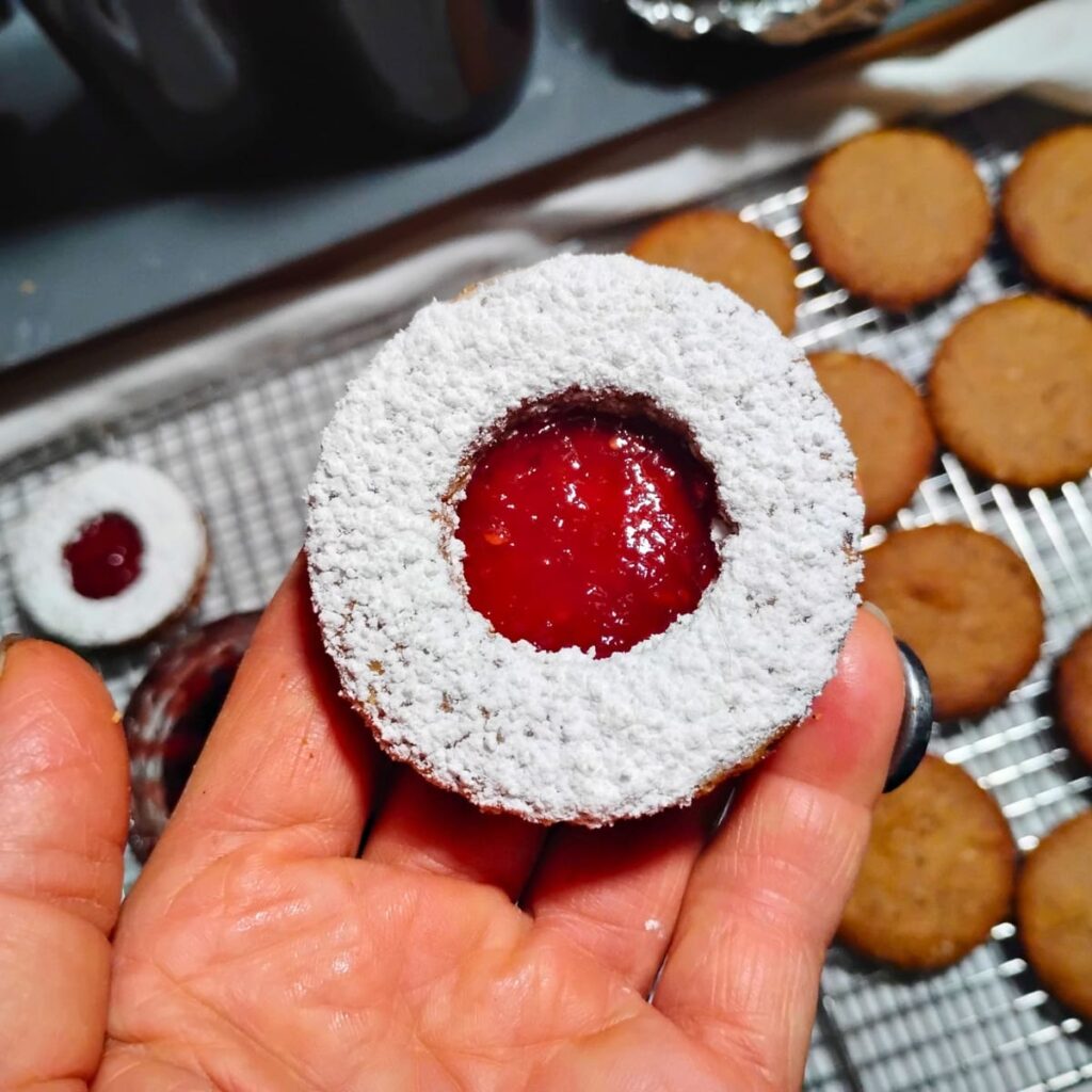 a person's hand holding up a finished cookie after placing the sugared ring top onto the jam with the linzer eye revealing the red raspberry jam. More cookies are below on a rack waiting to get filled.