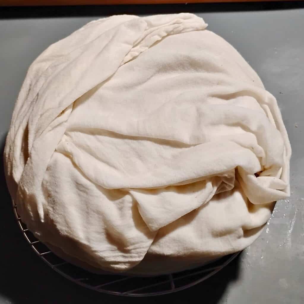 a loaf of bread wrapped in damp floursack towel on a wire rack.