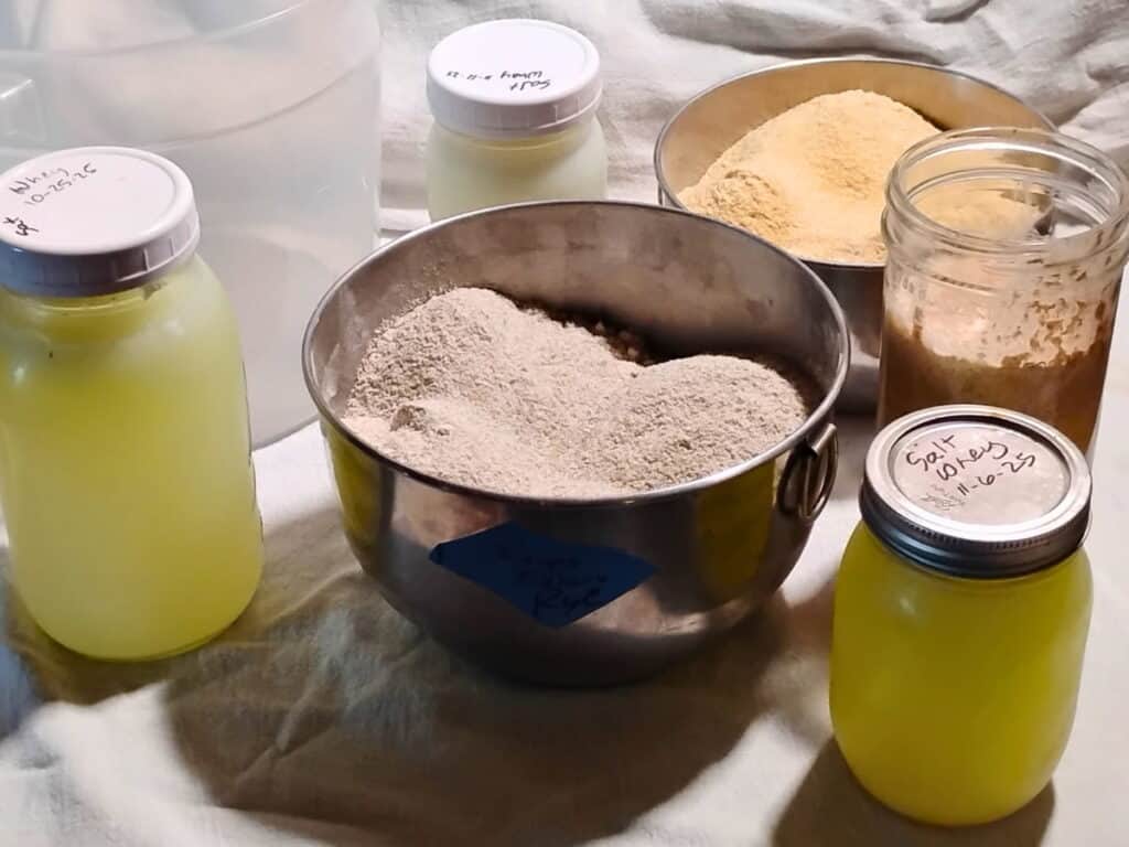ingredients for making the recipe. 3 jars of whey, a bowl with rye flour and kamut flour, and sourdough in a jar.