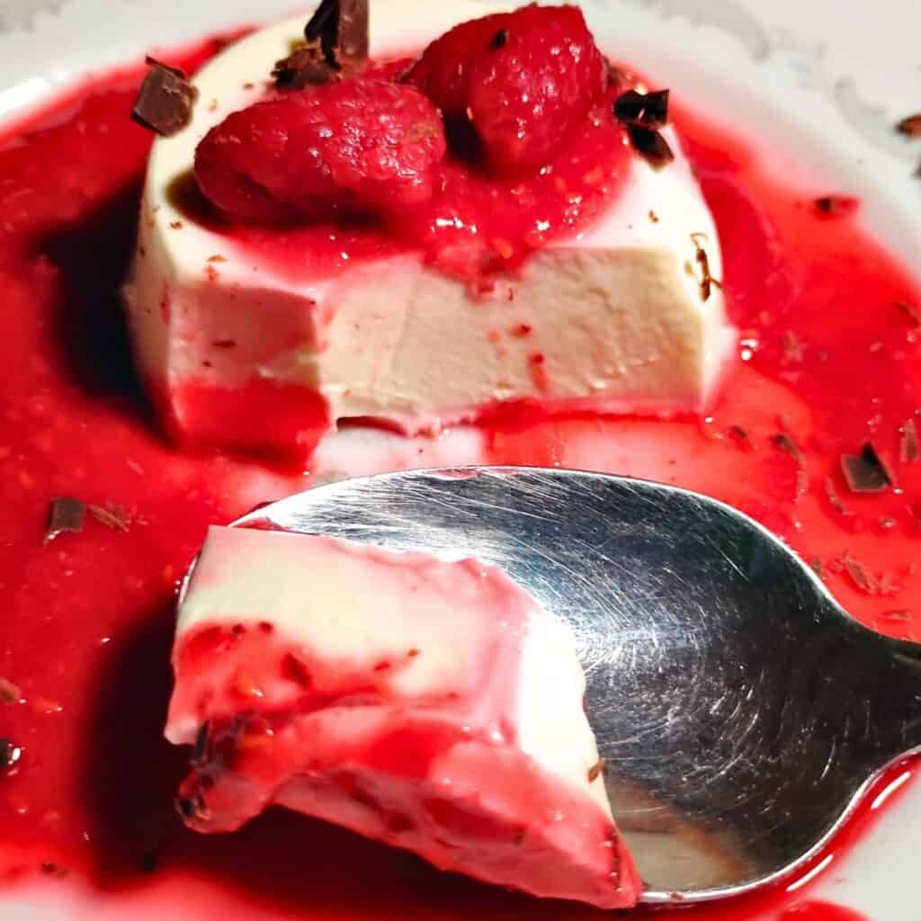 photo of a spoon taking a piece of panna cotta behind it that is covered with raspberries and surrounded by raspberry sauce on a plate.