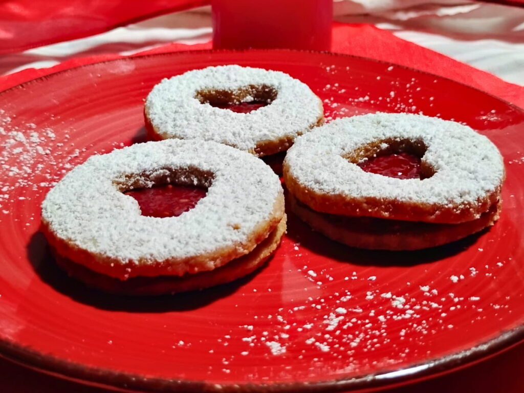 photo of 3 linzer cookies on a red plate. they are filled with raspberry jam and have heart shaped peekaboo cutouts.