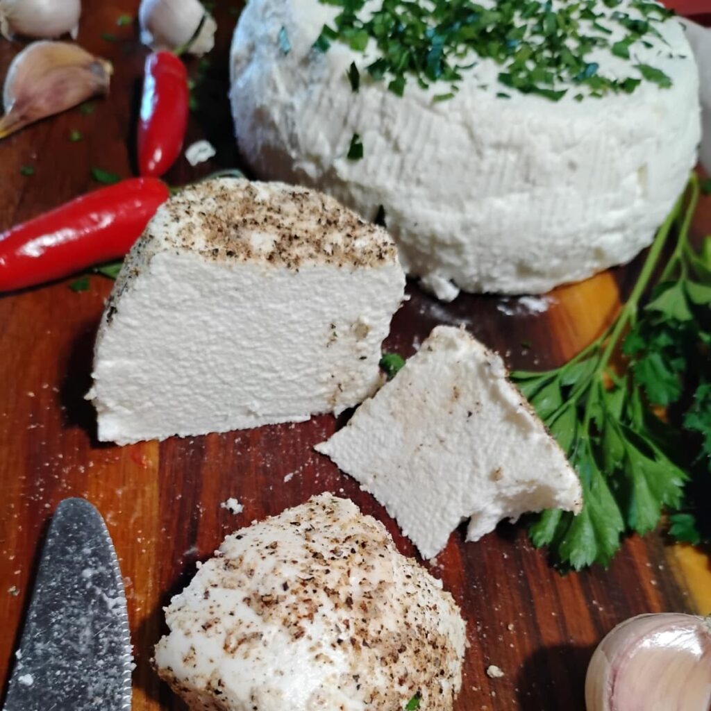 a wooden board with some chevre cheese chunks and a whole round of chevre that is covered with parsley behind it. 
