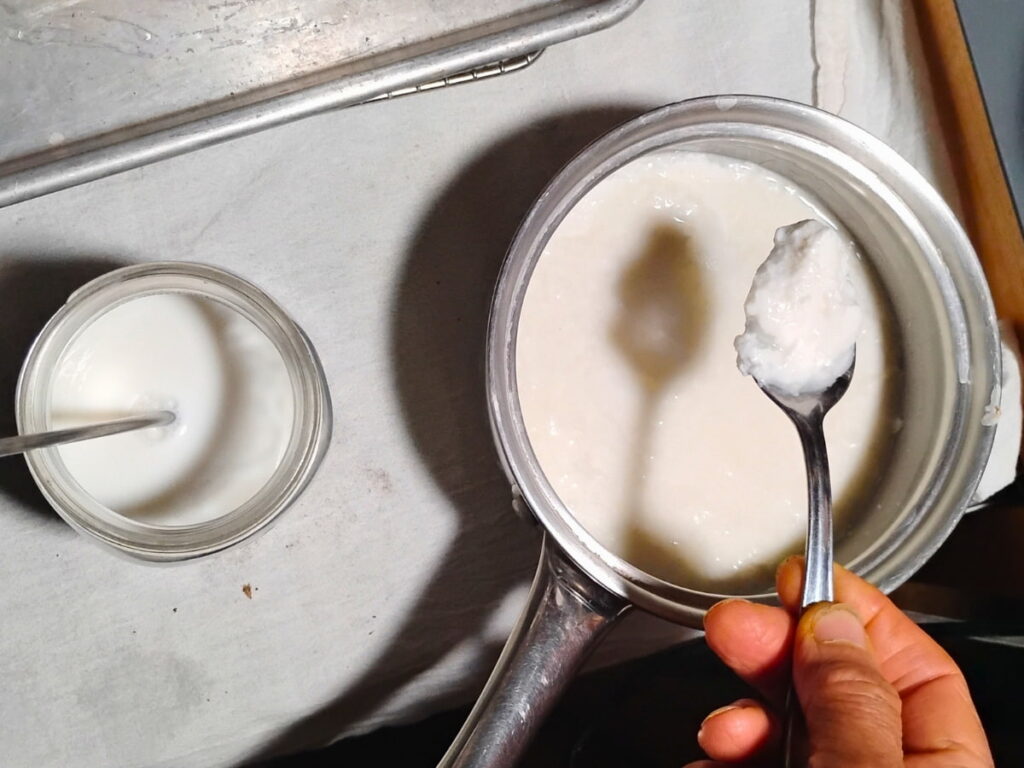 someone's hand is holding a spoon with a load of clabber that is taken from a pot below it and is getting transferred to a pint jar filled with warm milk next to it.