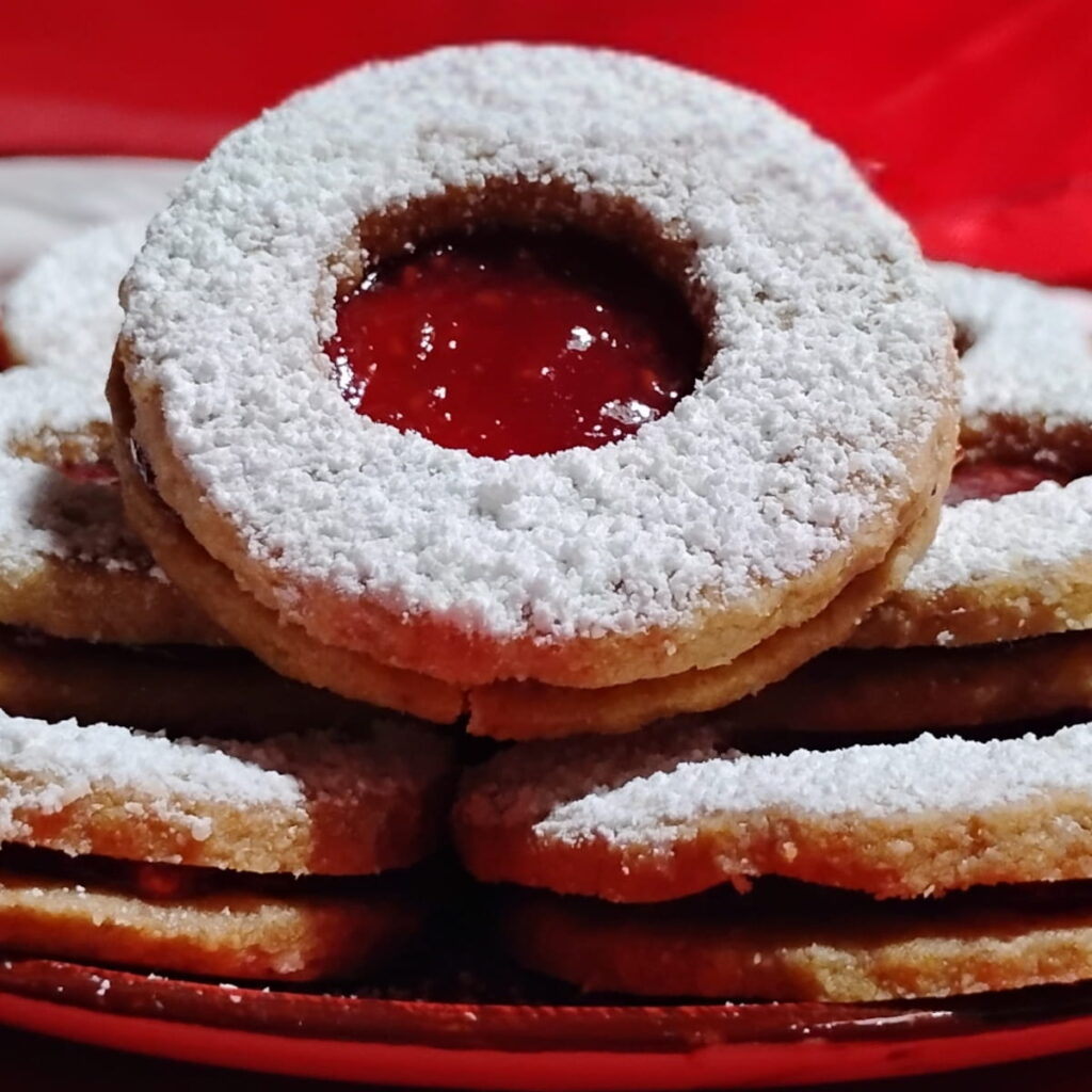 closeup photo of a linzer cookie with a red linzer eye, filled with raspberry jam. There are more cookies behind it on a plate.