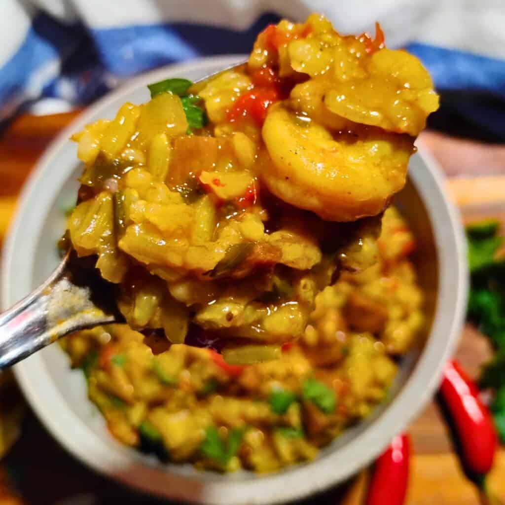 a spoon with a pile of ham jambalaya on it and a bowl that is full of jambalaya is below it on a table.