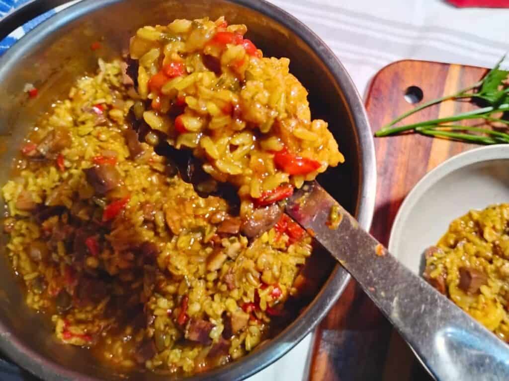 photo of a pot filled with ham jambalaya and a bowl with a few spoonfuls next to it. There is a spoon taking some ham jambalaya from the pot and transferring it to the bowl.