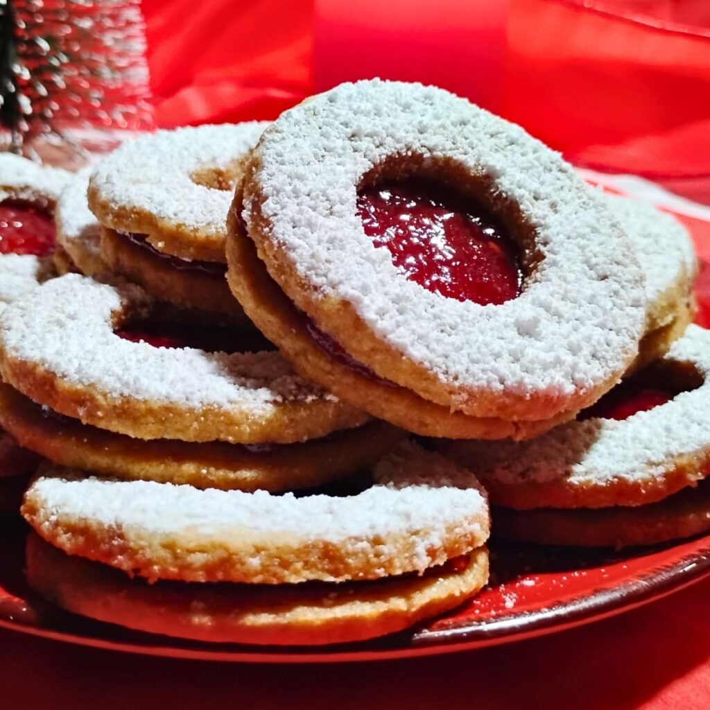 closeup photo of a plate piled up with linzer cookies that are filled with red jam. there is christmas decoration in the background.