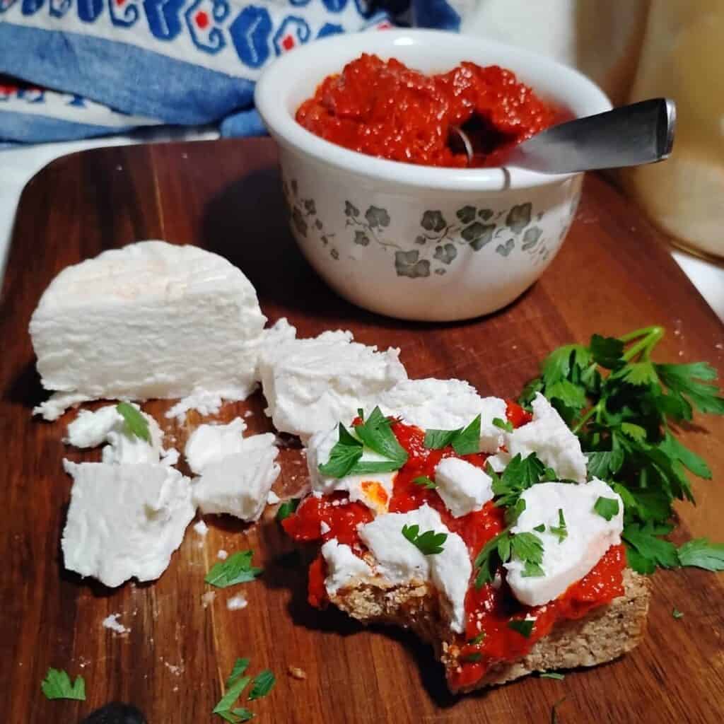 a piece of sourdough rye bread, topped with ajvar and soft feta cheese and some parsley. there is a small chunk of soft feta cheese and feta crumbles scattered next to it and a bowl filled with deep red ajvar behind it. 