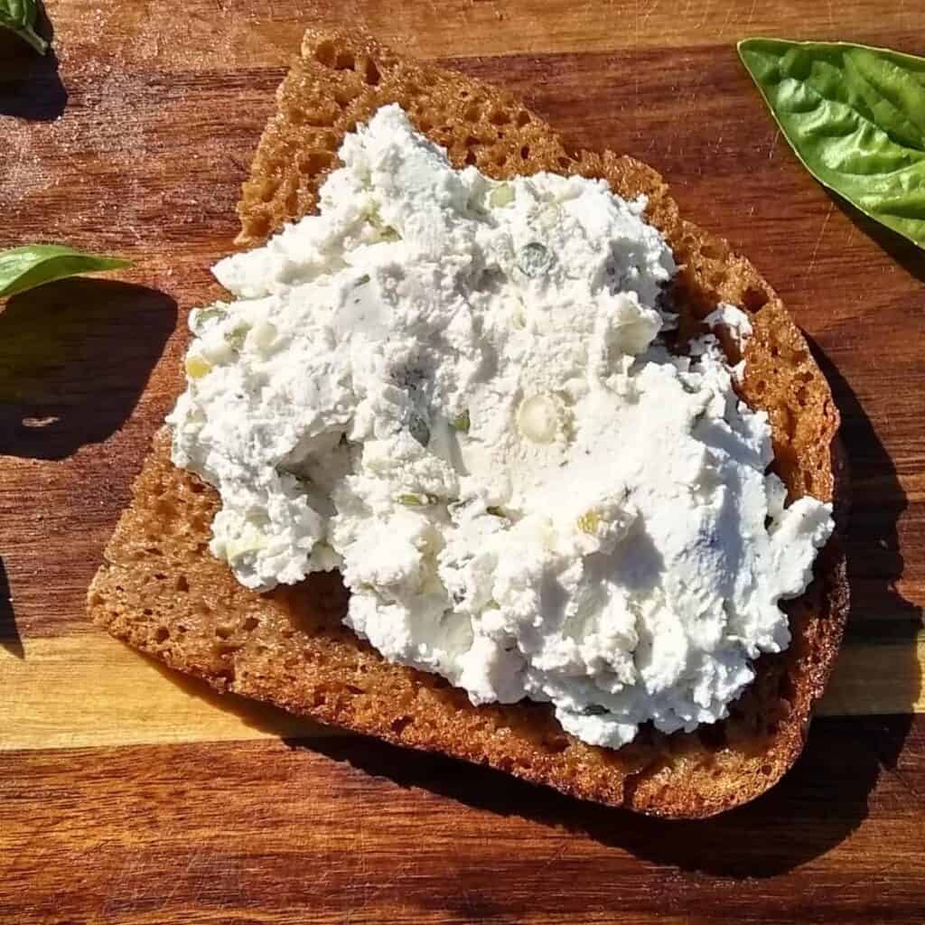 A slice of Kamut and Rye sourdough on a wooden board with creamy cheese that is mixed with herbs. 