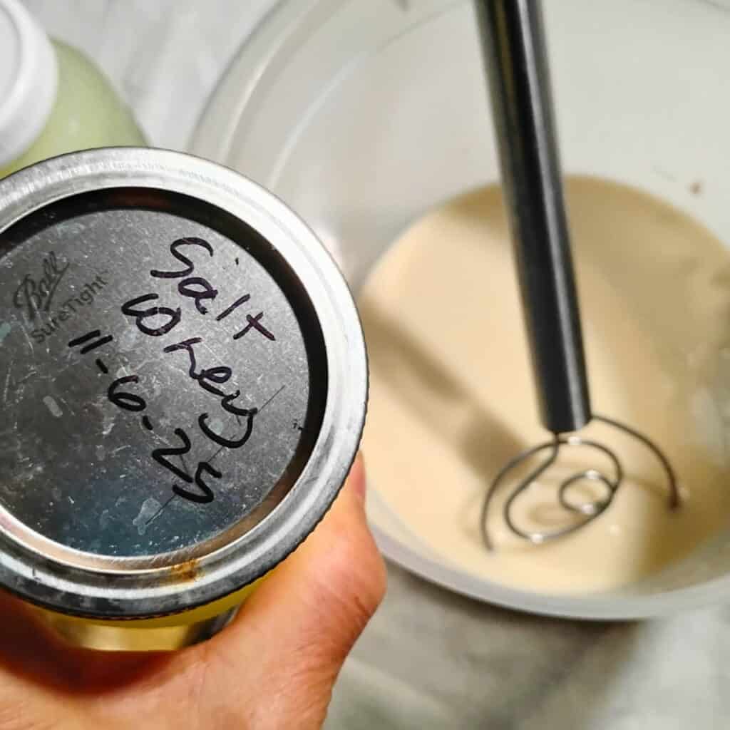 photo of a hand holding a jar filled with salted whey over a bowl with sourdough and a danish dough whisk. 