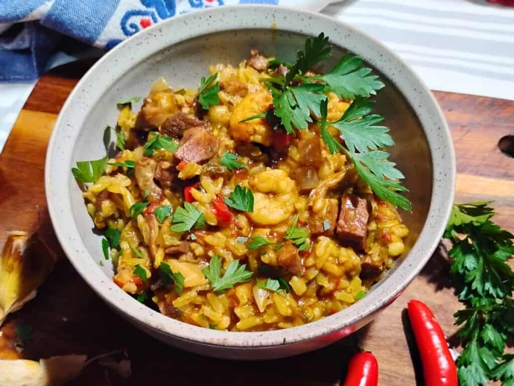 photo of a bowl filled with shrimp and ham jambalaya topped with some parsley. there are red serrano peppers, garlic, and parsley next to it on a cutting board.