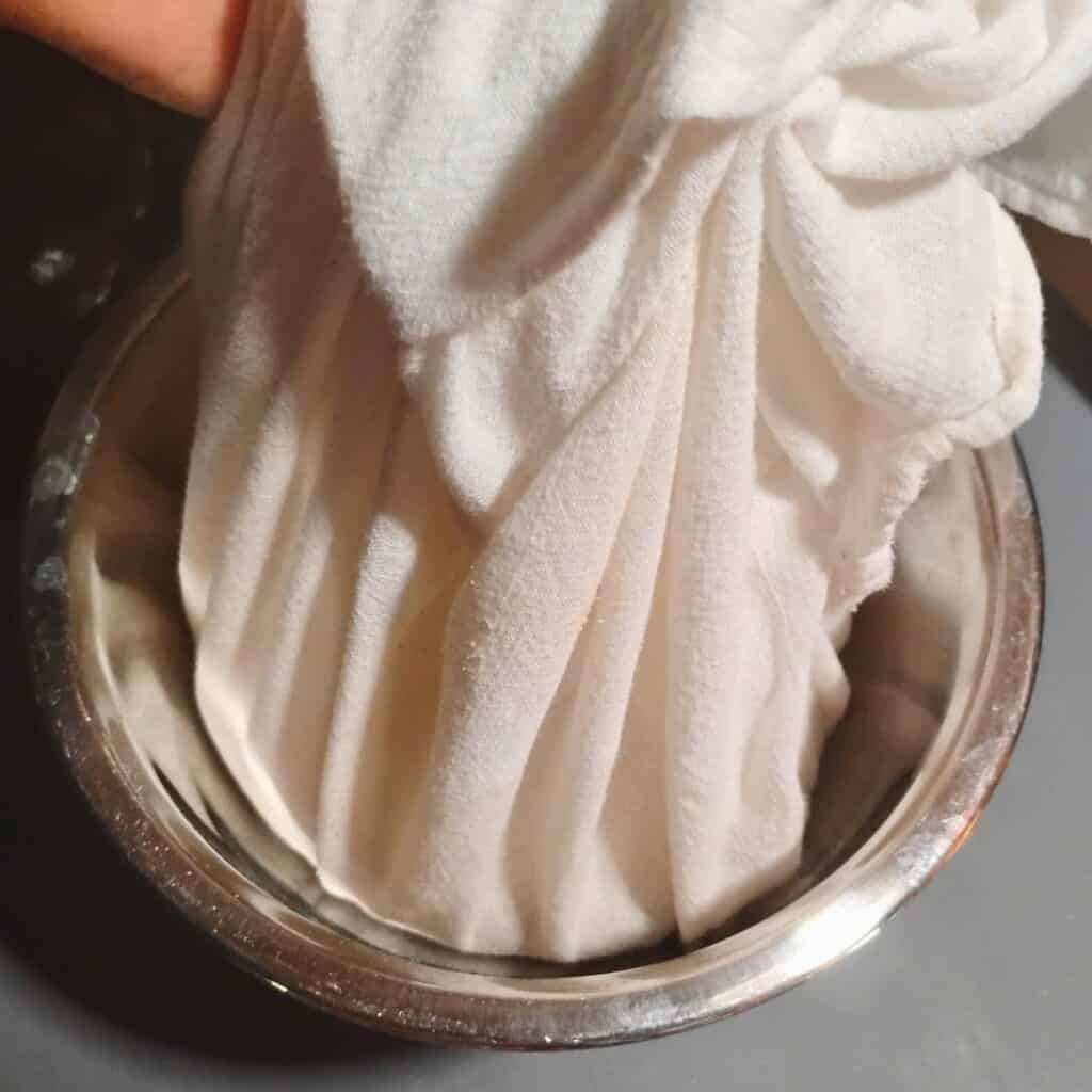 a hand holding the corners of a cloth with dough in it and lifting it into a bowl. 