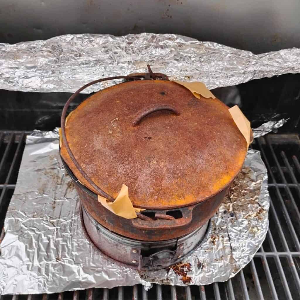 demonstration of grill baking setup. a aluminum foil at the bottom, a round cake ring to add height, a dutch oven with lid and parchment corners sticking out and aluminum foil in back of it to cover part of the back opening. 