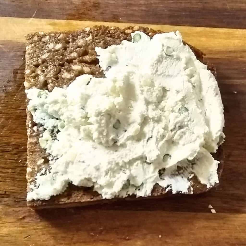 A slice of toasted seeded rye bread with a smear of herbed chevre cheese on top.