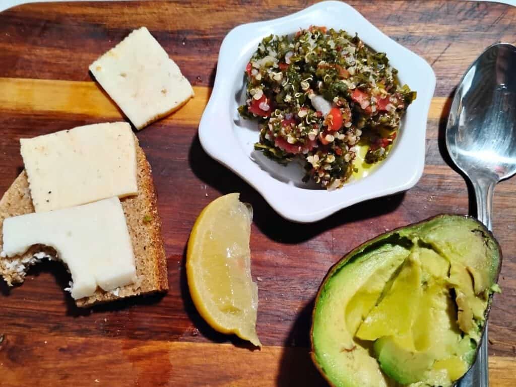 a spread of mezze with bel paese cheese on a slice of bread, an avocado half and a small plate with fermente tabouli salad. Arranged on a wooden board.