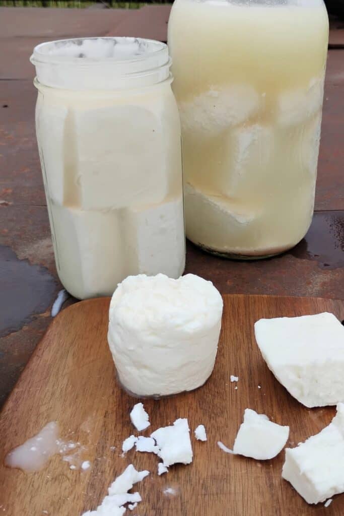 two tall jugs filled with feta cheese. one has harder sirene feta in it, the other has soft feta in it. iIn front are a few chunks of feta. cheese on a cutting board.