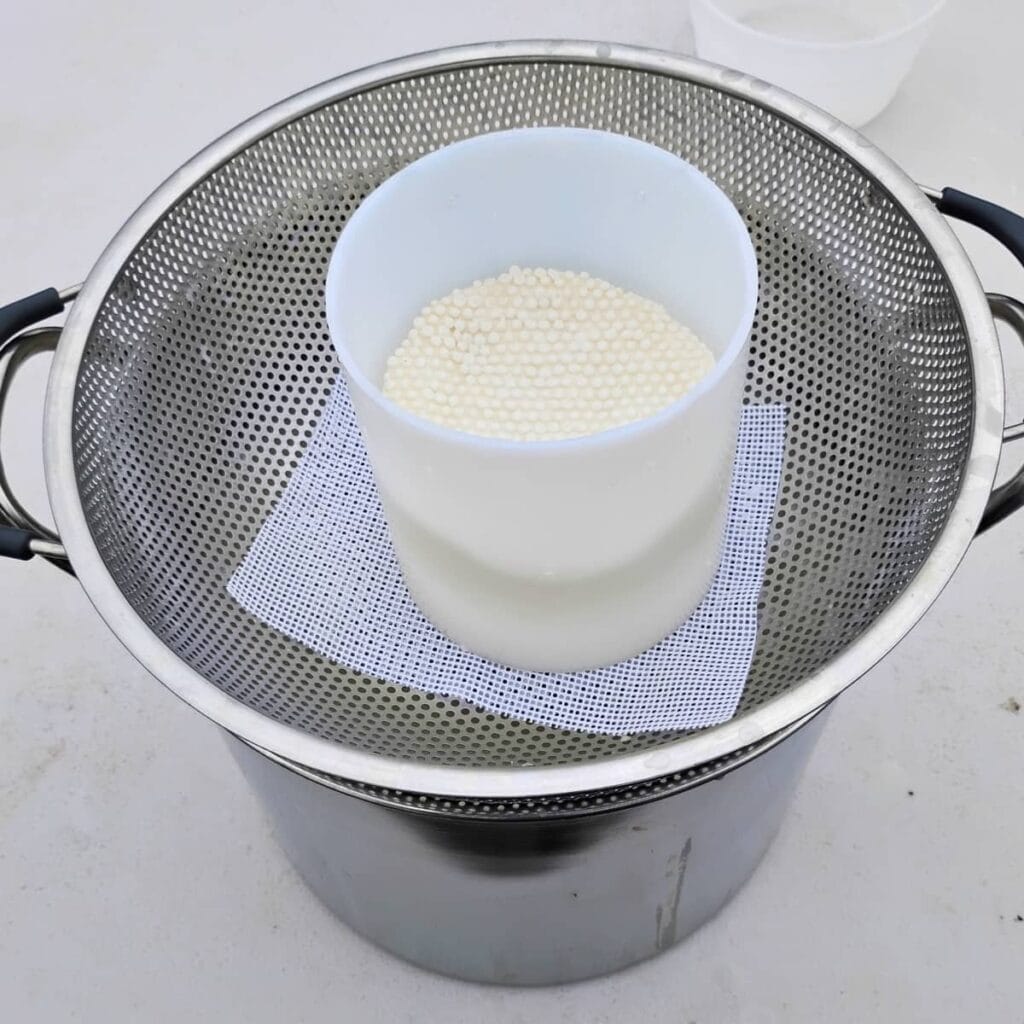 photo of a cheese draining setup with a pot, colander, drainage cloth, cheese mold and curd inside.