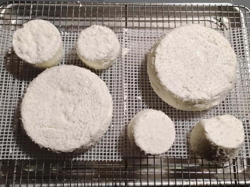 a feta cheese draining station made up of a baking sheet with a lasagna pan in it and a wire rack over it. On it are a couple of cheese mats and chunks of feta cheese draining on them.