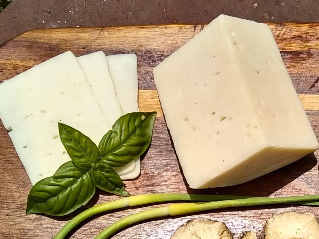 3-year old Bel paese cheese chunk on a cutting board and few slices next to it with some basil and garlic scapes.