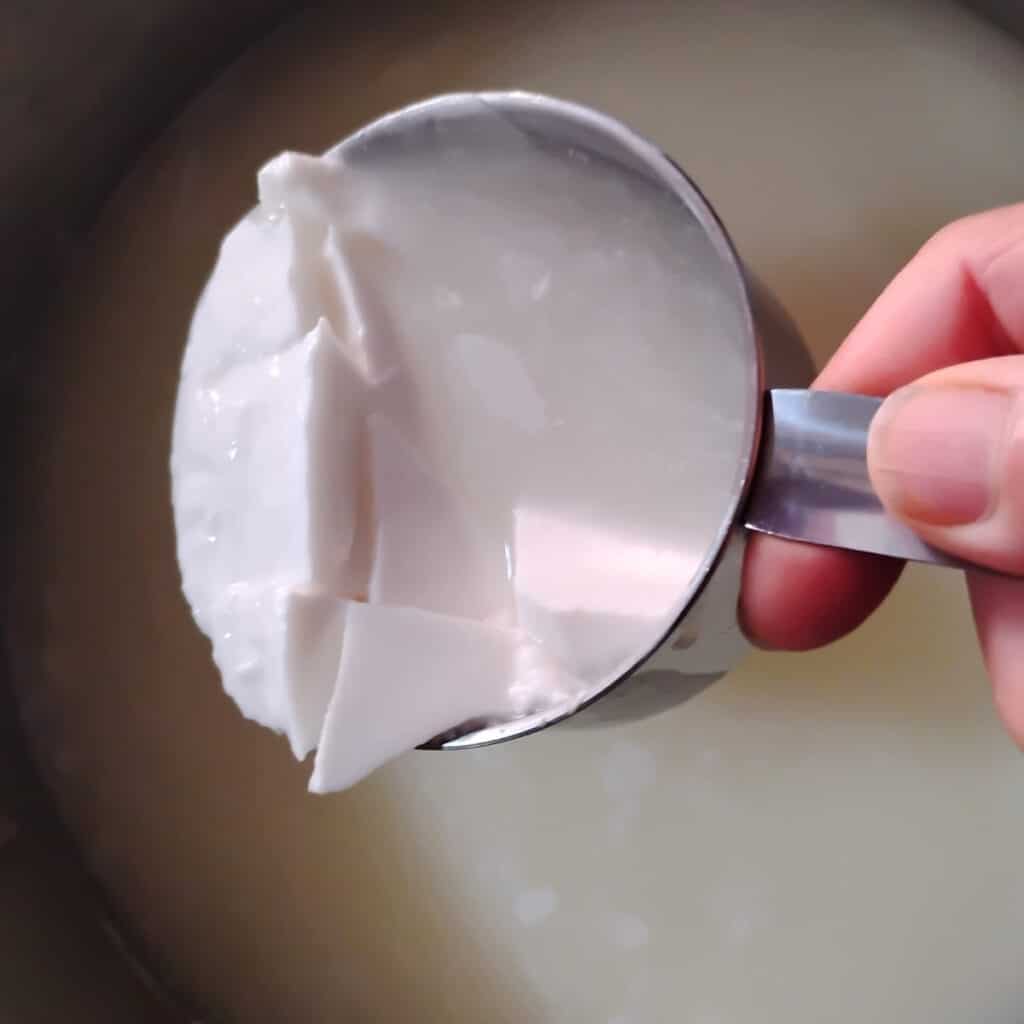 a measuring cup in a person's hand scooping cheese curd chunks out of a pot,