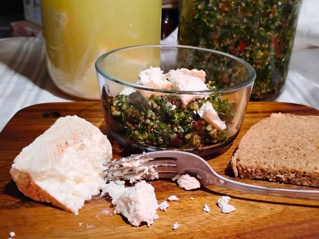 photo of a small glass bowl with quinoa salad and crumbled feta cheese. there is more feta cheese beside it on a wooden board with a fork. A slice of rye bread is on the other side of it. behind are two jars, one with feta cheese chunks and brine, the other with more quinoa tabbouleh salad. 