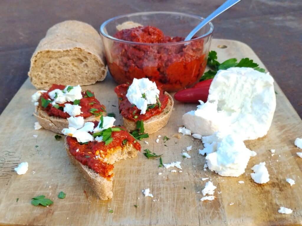 photo shows a piece of bread and a bowl filled with ajvar dip. there are a few pieces of bread with ajvar and feta cheese on top. there is a chunk of feta cheese also.