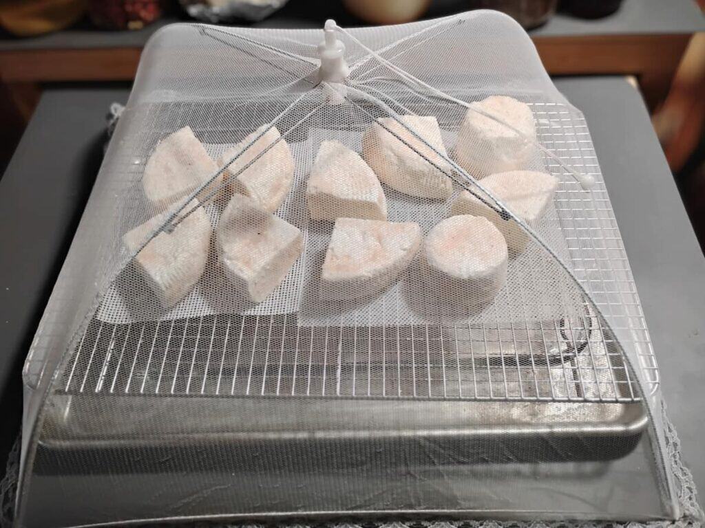 a feta cheese draining station made up of a baking sheet with a lasagna pan in it and a wire rack over it. On it are a couple of cheese mats and chunks of feta cheese draining on them. All of it is covered with a netted food cover umbrella.