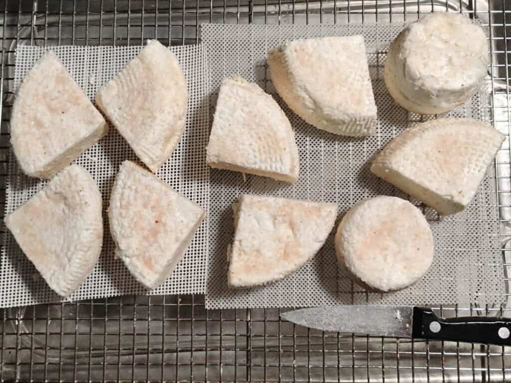 a feta cheese draining station made up of a baking sheet with a lasagna pan in it and a wire rack over it. On it are a couple of cheese mats and chunks of feta cheese draining on them. The feta chunks are covered with salt.