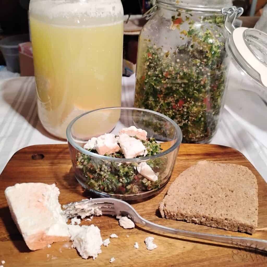 photo of two jars in the back with tabouli and brined feta cheese. in front is a small bowl with fermented tabouli and feta cheese chunks and a slice of bread on a cutting board.