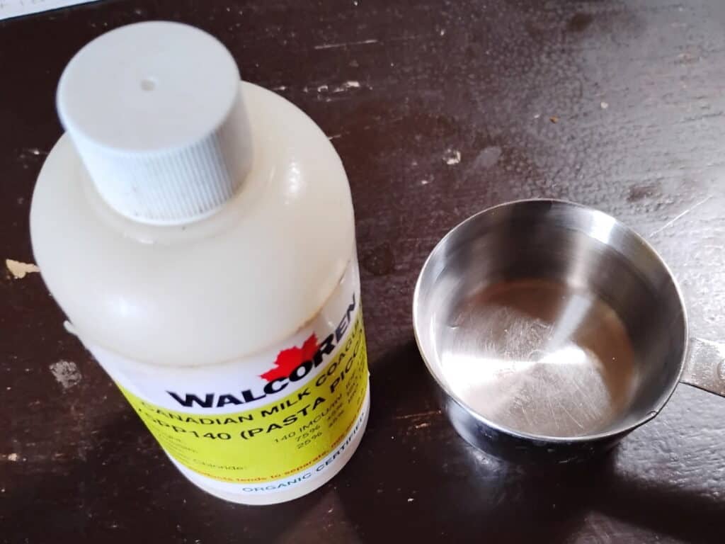 photo of a bottle of rennet and a measuring cup with some water next to it.