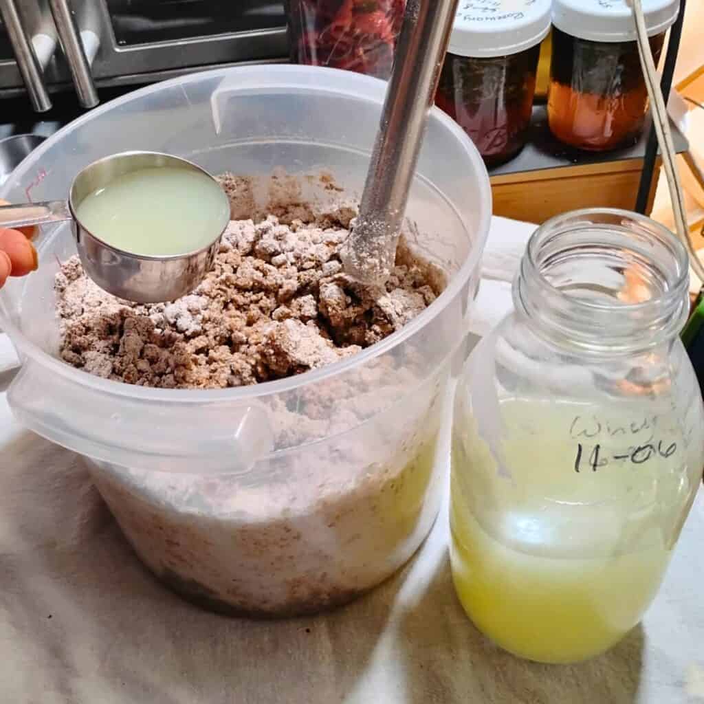 whey is getting added to a container filled with sourdough rye bread ingredients. there is a bottle of whey standing next to the bread dough bucket.