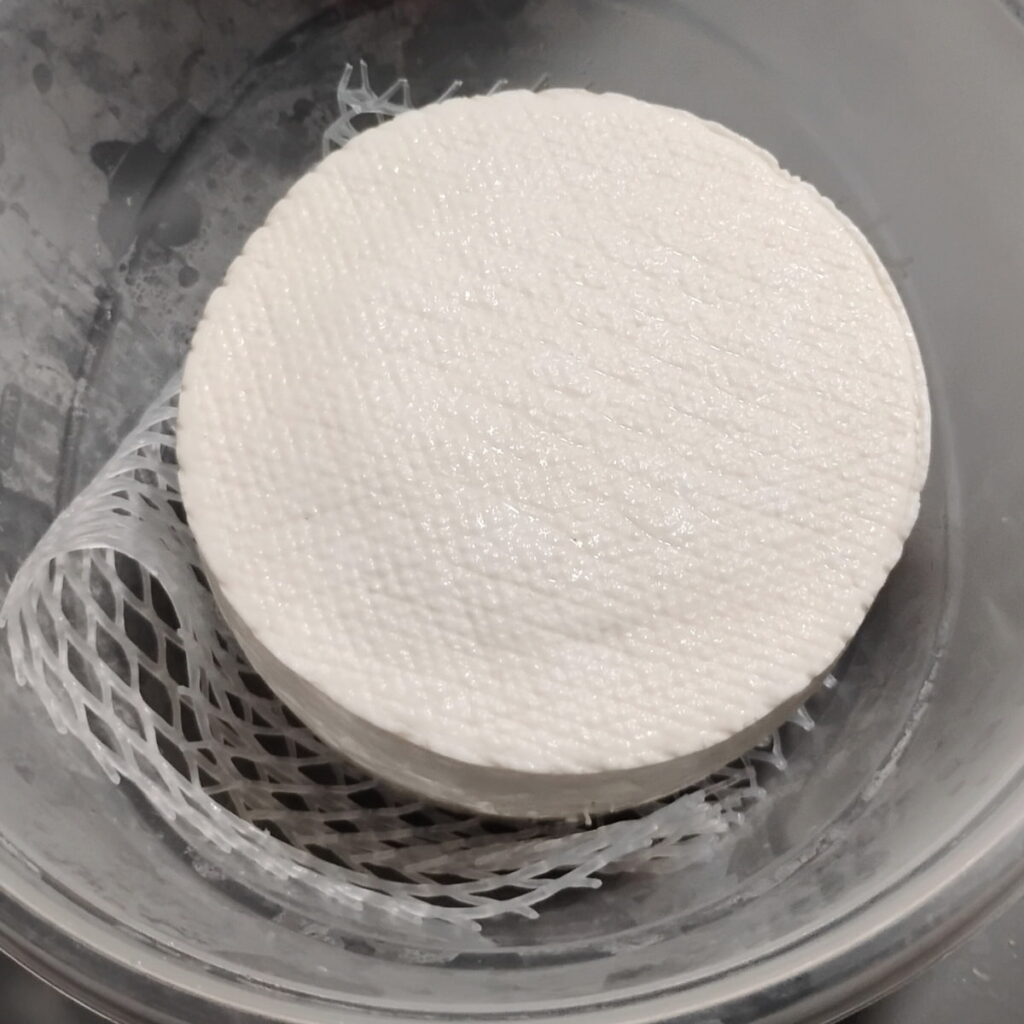 a loaf of bel paese cheese inside a plastic bowl with a piece of silicone mesh, ready to go into the fridge for ripening.