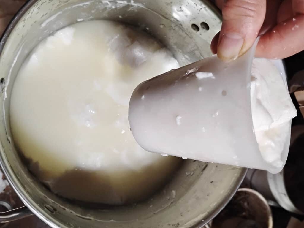 a person's hand is holding a small chevre form with cheese curds over pot.
