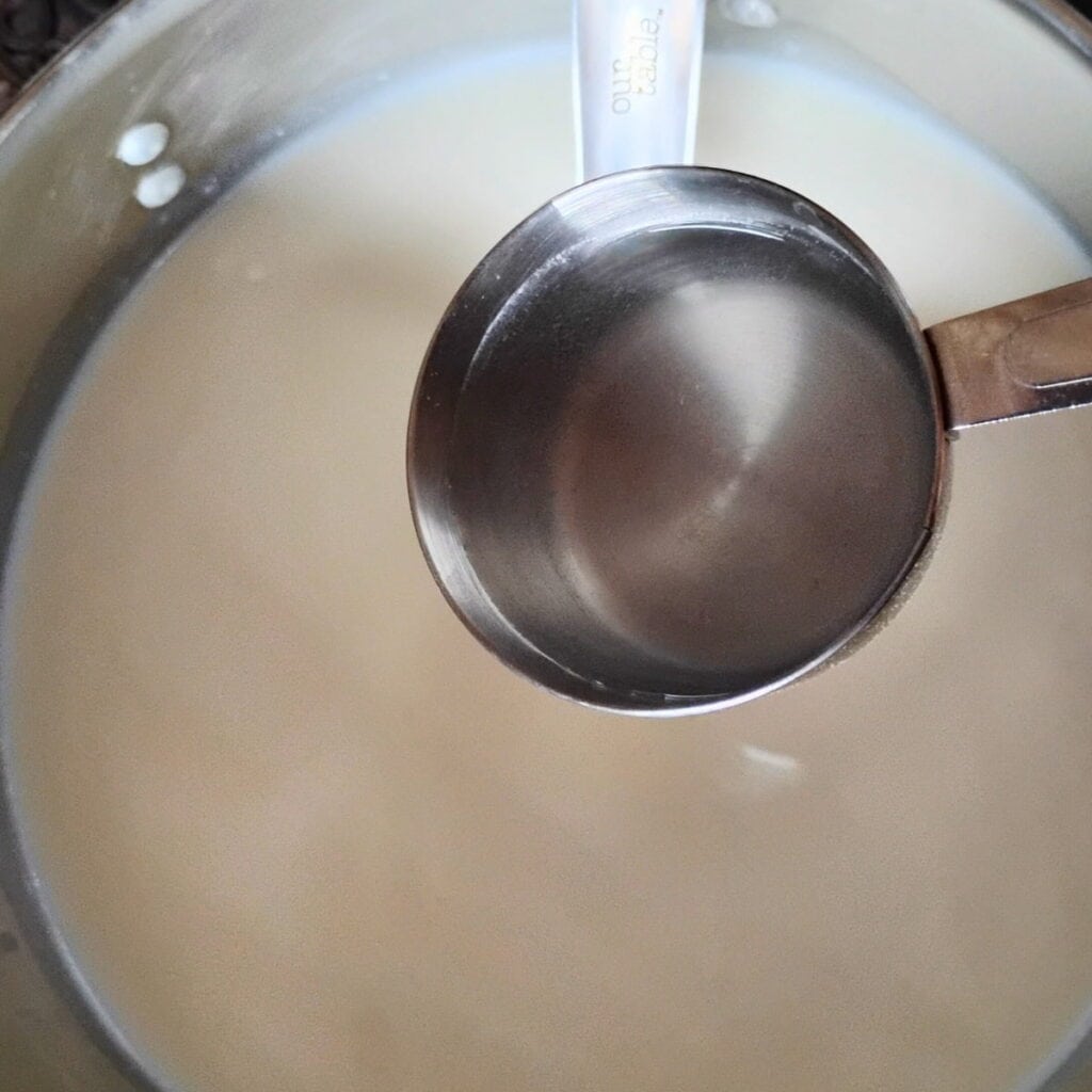A hand is holding a measuring cup with diluted rennet to be added to the milk in a pot.