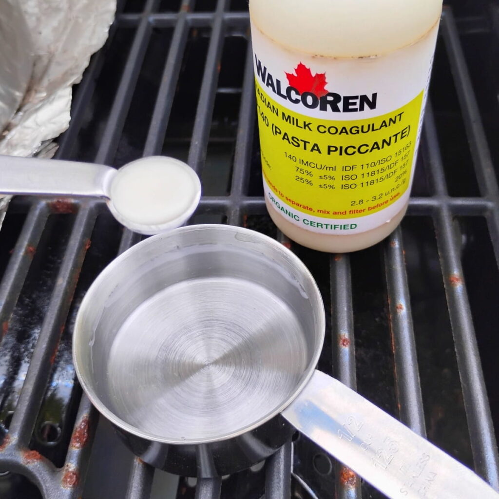 a bottle of organic kid rennet by walcoren, a small measuring cup with water and a measuring spoon filled with rennet to be mixed with the water.