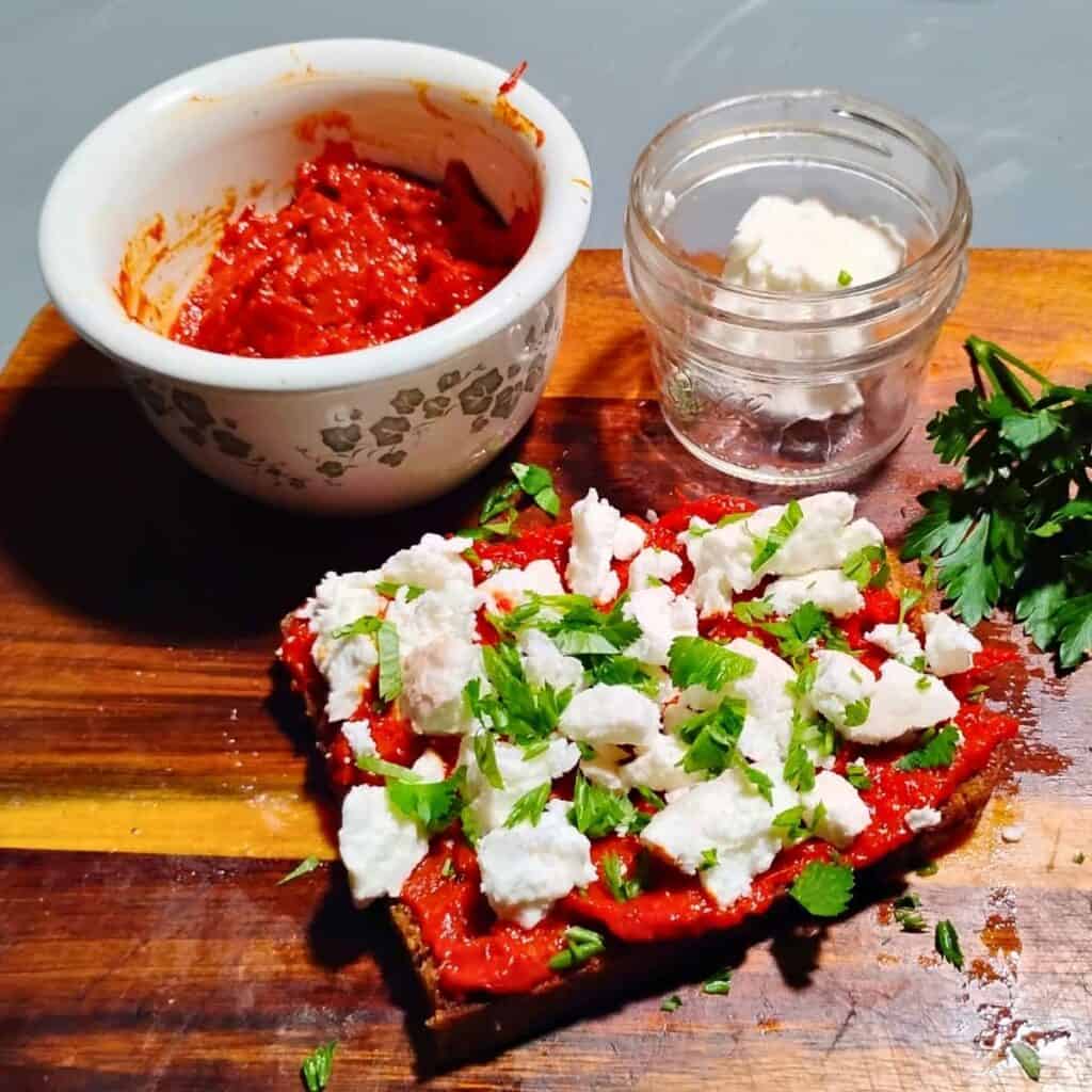 photo shows a jar filled with ajvar and a smaller jar with some feta cheese in it. in front is a slice of bread topped with ajvar spread and feta cheese chunks and parsley.