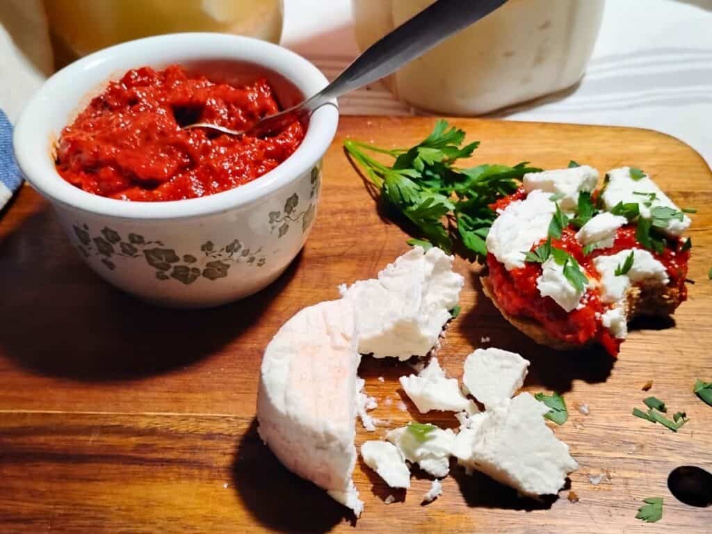 photo shows a couple of jars filled with brined cheese in the background. in front is a bowl filled with ajvar dip and a piece of bread with ajvar and feta chunks on it. There are more feta cheese chunks in the foreground.