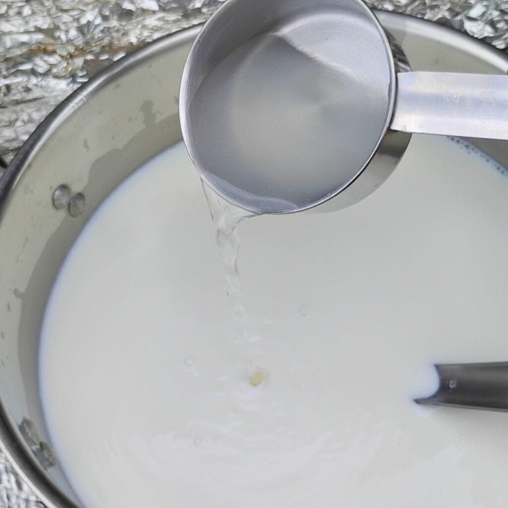 pouring a measuring cup full of water and rennet into the pot of milk.