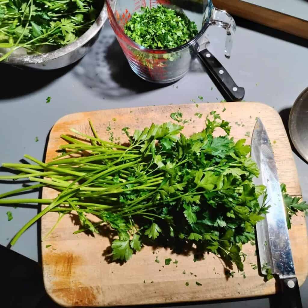 parsley on a cutting board with a knife and some chopped parsley in a dish behind it. 