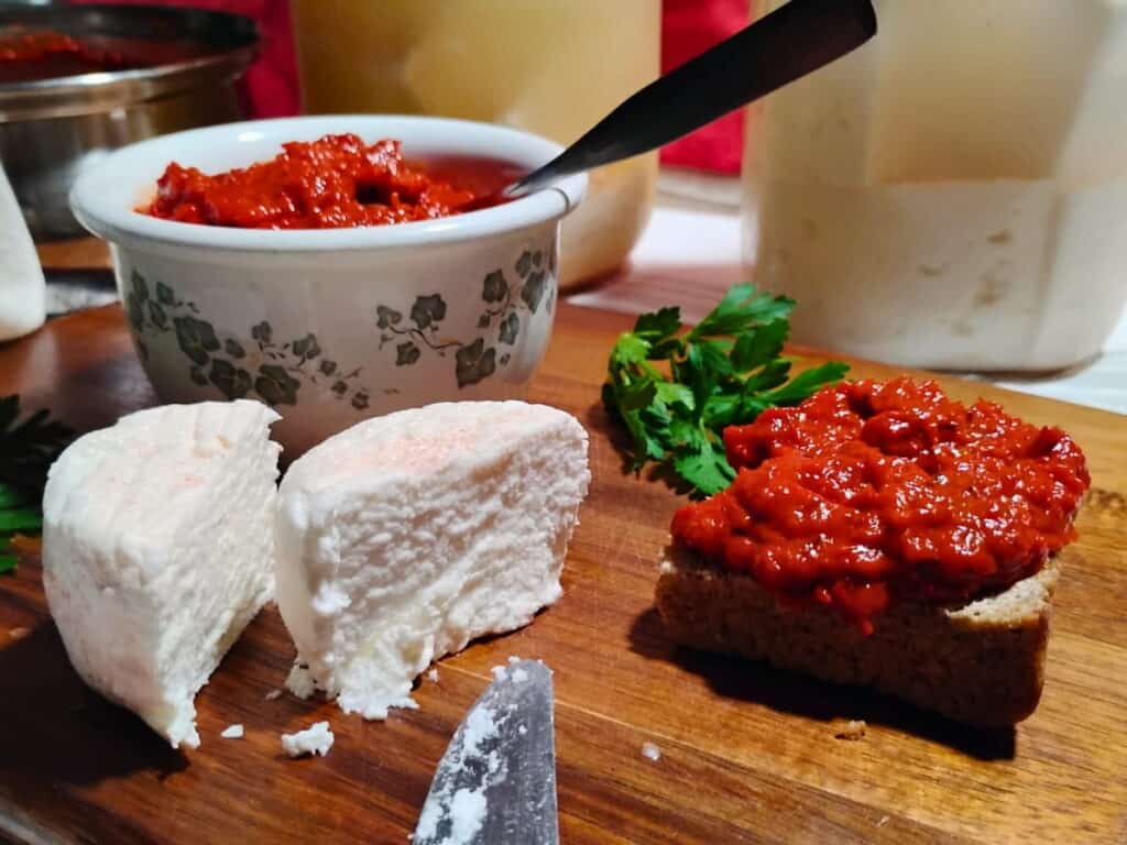 photo of a bowl filled with ajvar in the back and some jars filled with brine and feta chunks. in the forground is a chunk of feta cheese that has been cut in half and a piece of bread with some ajvar spread on it.