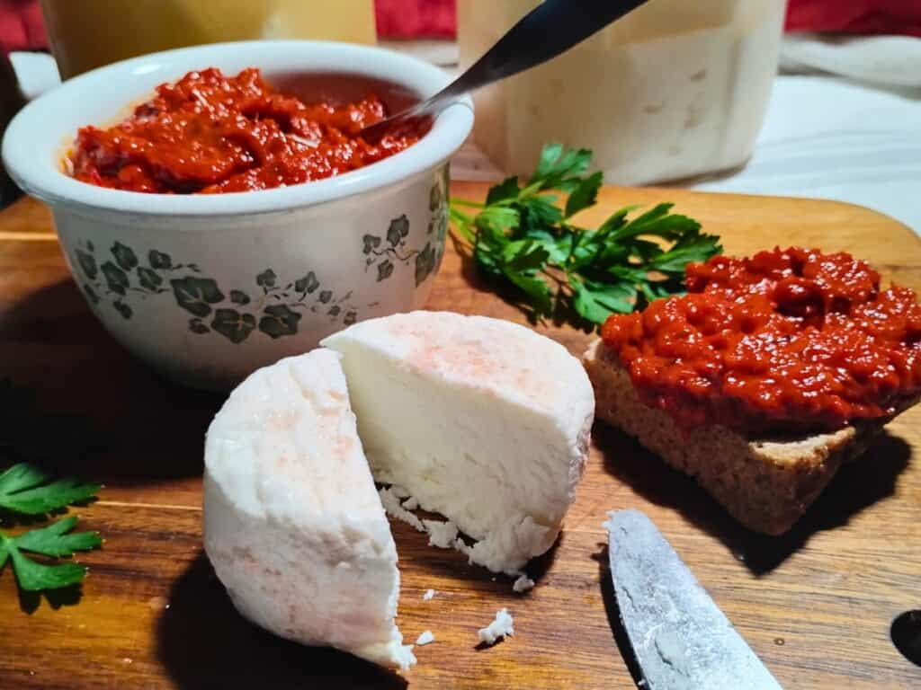 photo of a chunk of feat cheese that is cut in half. A bowl of ajvar is sitting behind it and a piece of bread with some ajvar is next to it.