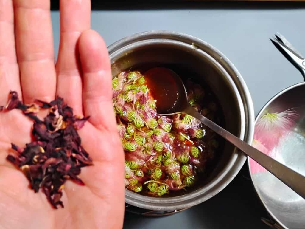 mimosa flowers in syrup in a pot and a hand holding some hibiscus flowers to add. 